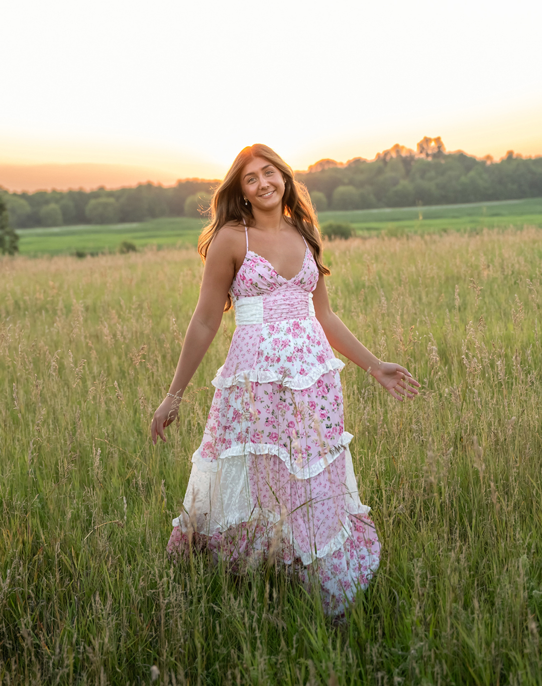 High school senior girl standing in tall grass at sunset wearing a flowing floral dress in a natural field setting.